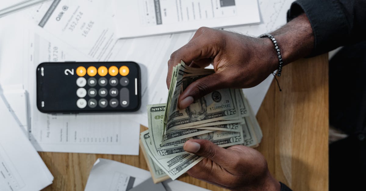 Person counting dollar bills over documents with a smartphone calculator on the desk.