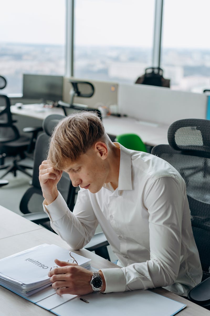 Young man in a modern office looking stressed with paperwork, symbolizing work pressure.